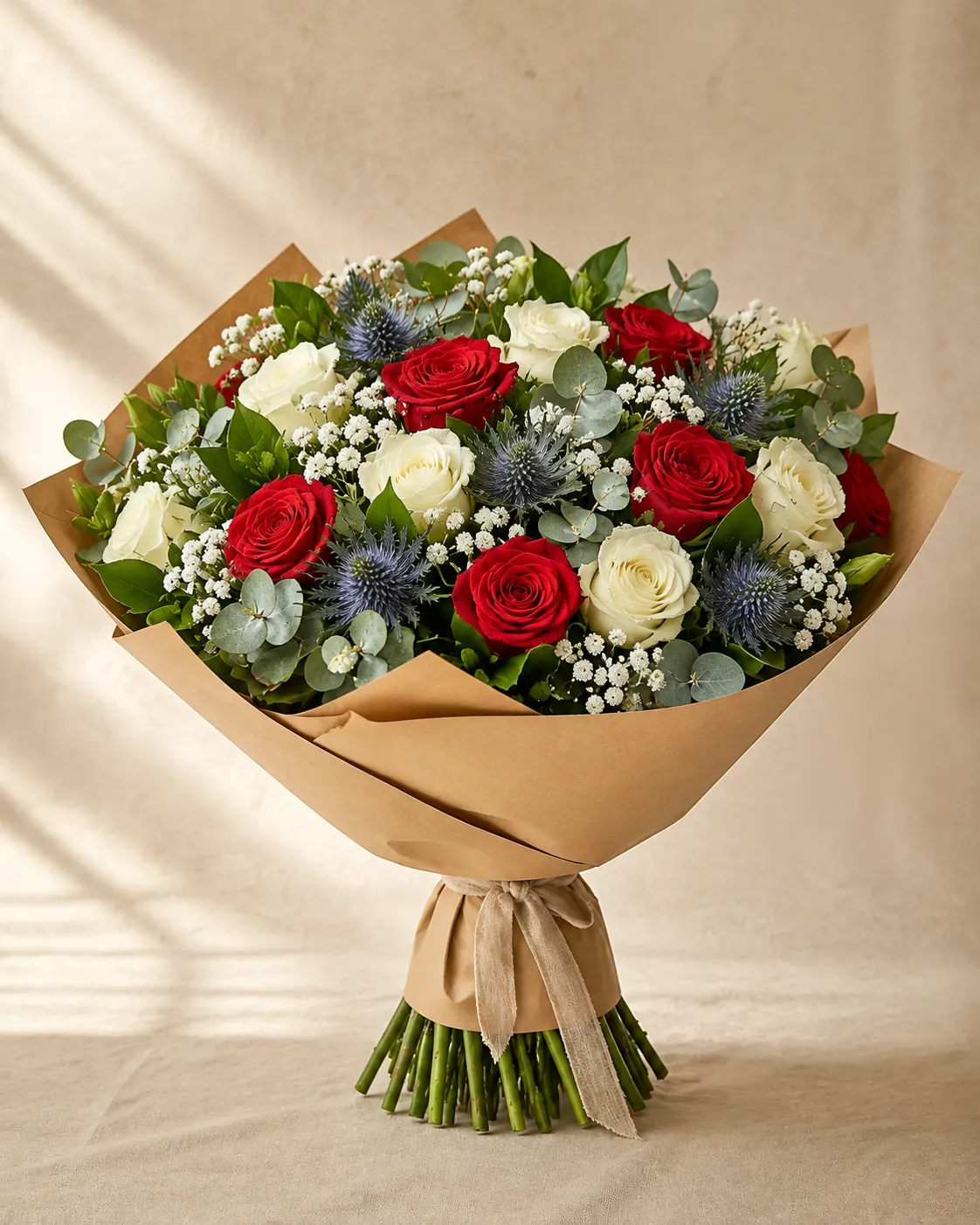Bouquet de roses rouges et blanches, chardons bleus, gypsophile et eucalyptus dans un papier kraft noué d’un ruban de lin